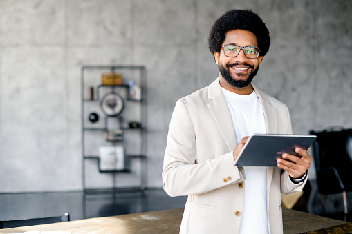 The young office professional holding a tablet, standing against an industrial backdrop, a modern, creative work environment. His smile suggest a friendly approach to business
