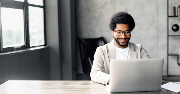A cheerful Brazilian businessman in a smart casual beige suit in loft office using laptop Stylish professional is fully engaged with his work on a laptop, reflecting a scene of modern business efficiency and concentration. His smart casual outfit suggests relaxed yet productive atmosphere black man stock pictures, royalty-free photos & images