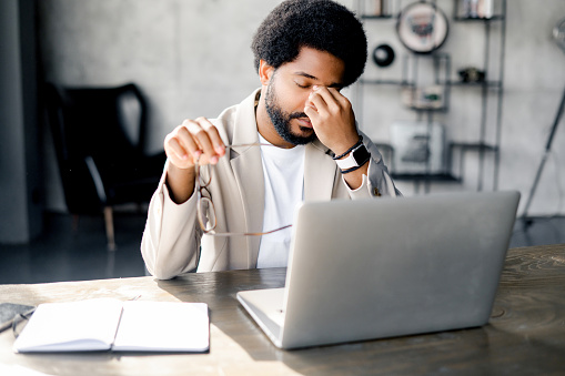 Modern businessman rubs eyes in fatigue or exasperation while working on his laptop, a depiction of the demanding nature of contemporary work life. Male office employee feels burnout and overworked