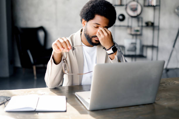 A modern businessman rubs his eyes in fatigue or exasperation while working on his laptop Modern businessman rubs eyes in fatigue or exasperation while working on his laptop, a depiction of the demanding nature of contemporary work life. Male office employee feels burnout and overworked managing stress stock pictures, royalty-free photos & images