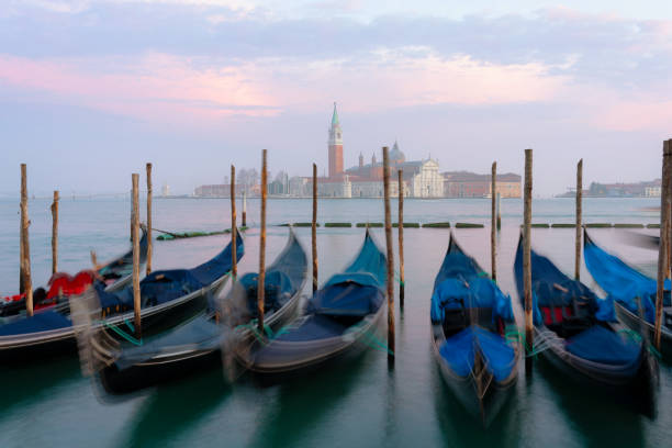 A beautiful sunset illuminates a line of gondolas tied up on the side of the Grand Canal in Venice. Across the lagoon we see the iconic shape of the Church of San Giorgio Maggiore.