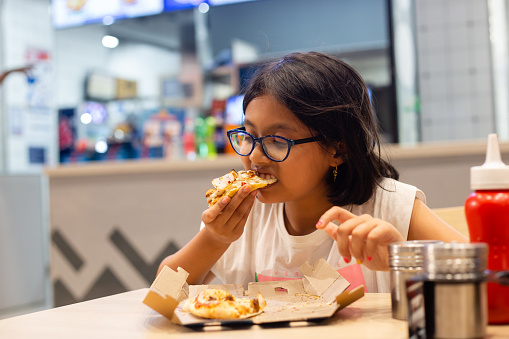 A petty Indian girl child eating pizza sitting at a restaurant table at night