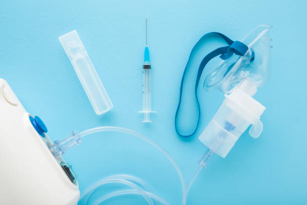 istock Compressor nebulizer, plastic ampoule with liquid, syringe and transparent mask with container for drug breathing on light blue table background. Closeup. Medical device for inhalation. Top down view. 2022830501