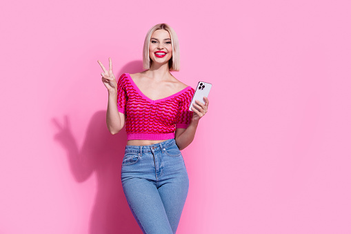 Photo of toothy beaming cheerful woman with bob hairstyle dressed knit top showing v-sign hold smartphone isolated on pink background.