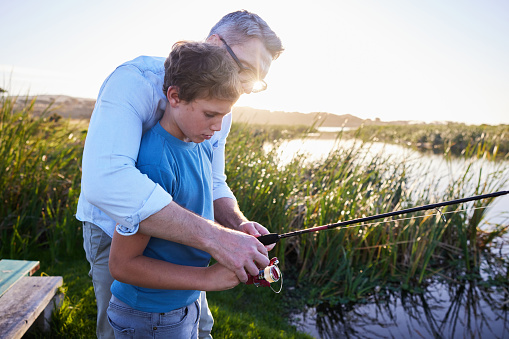 Loving father teaching his smiling young son how to fish with a rod during a vacation together at a scenic lake on a summer afternoon