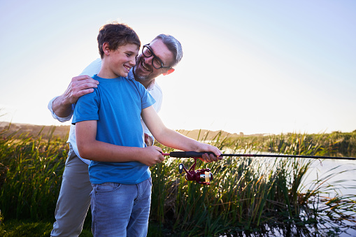 Loving father teaching his smiling young son how to fish with a rod during a vacation at a scenic lake on a sunny summer afternoon