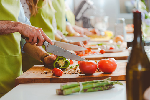 Group of people wearing aprons taking part in cooking class, preparing food, slicing vegetables. Close up of hands, unrecognizable people.