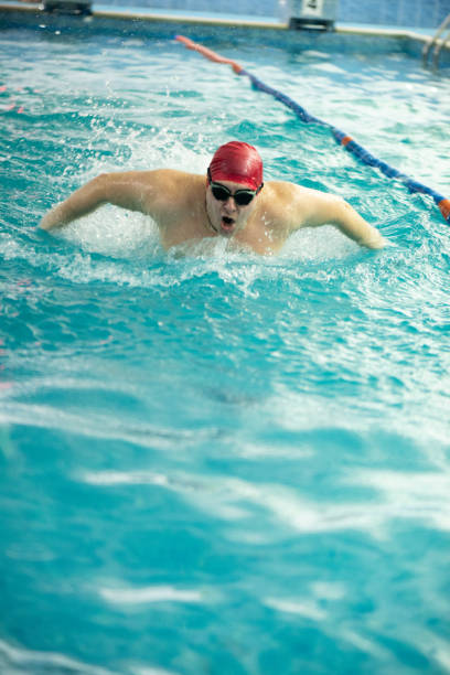 Dynamic shot of a young adult male swimming in a indoors pool wearing red cap and swimming goggles