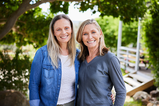 Woman and her mature mother smiling while standing arm in arm together outside in a back yard at home in summer