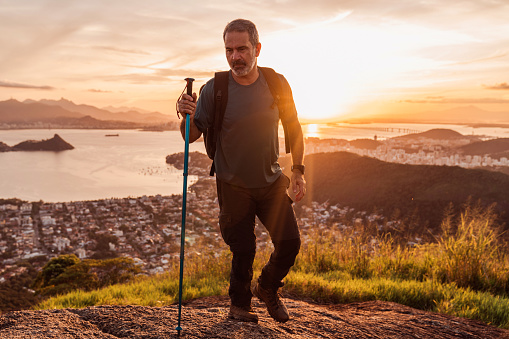 A solitary hiker stands at the summit of a hill, gazing out at the stunning landscape of  the city during the golden hour. The sun sets in the distance, casting a warm glow over the city and its iconic mountainous coastline.