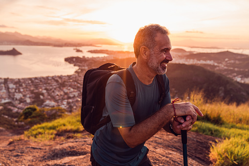 A solitary hiker stands at the summit of a hill, gazing out at the stunning landscape of  the city during the golden hour. The sun sets in the distance, casting a warm glow over the city and its iconic mountainous coastline.