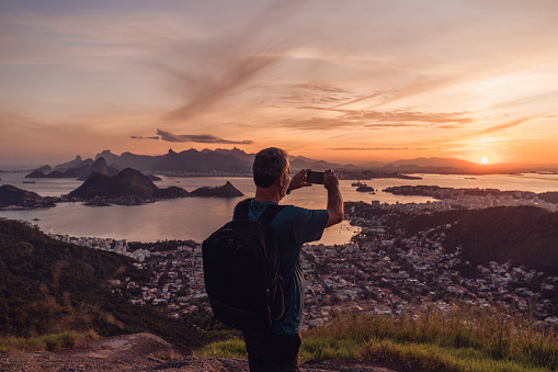 A man stands on a hilltop with a trekking pole, gazing at the breathtaking landscapes below him and taking photos with his smartphone