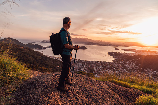 A solitary hiker stands at the summit of a hill, gazing out at the stunning landscape of  the city during the golden hour. The sun sets in the distance, casting a warm glow over the city and its iconic mountainous coastline.