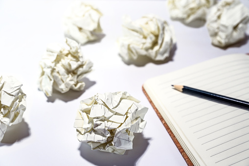 Discarded crumpled paper balls around an empty notebook with a pencil, concept for creative writing, development process, business frustration and stress, white table, copy space, selected focus