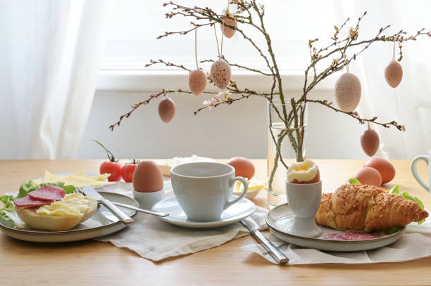 istock Easter breakfast with egg, bun, coffee, croissant, cheese and sausage, decorated with spring branches and hanging easter eggs in a glass vase on a table at the window, copy space 2022919051