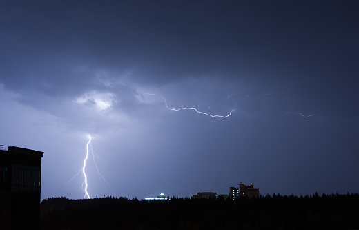 The dramatic scene of a lightning storm occurring above a cityscape at night. The bright, jagged lines of lightning illuminate the dark sky, contrasting against the city lights below. Its a powerful depiction of natures force amidst urban life.