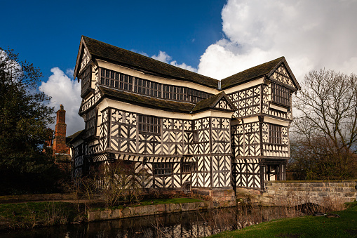 Little Moreton Hall, Cheshire, England, United Kingdom - February 26, 2011: The Black and white wooden framed Tudor house stands amid trees and is reflected in the water of the moat against a blue sky with large white clouds.