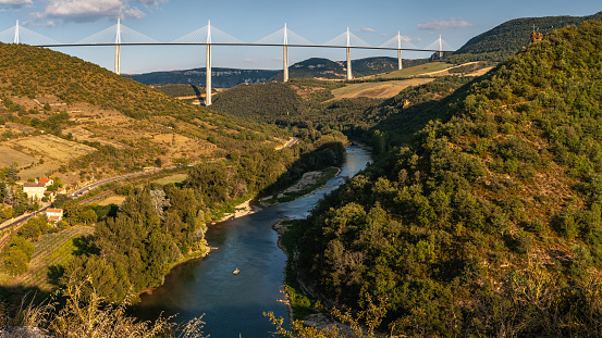 Millau, Aveyron, France - August 08, 2006: Panoramic view of the Millau Viaduct suspension bridge , co-designed by Norman Foster, set in a green summer landscape of rolling hills and crossing the River Tarn on which sails a single canoe.