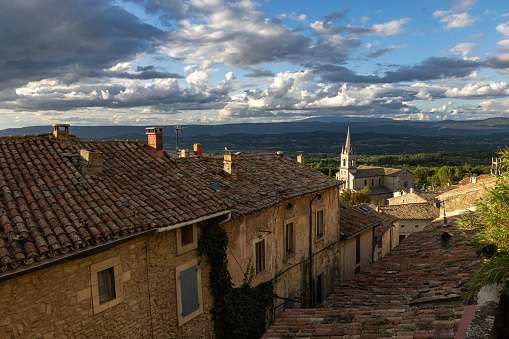 Ancient tiled roofs of the village of Bonnieux  and a distant church and in the very far distance Mount Ventoux covered with dramatic clouds in a blue sky