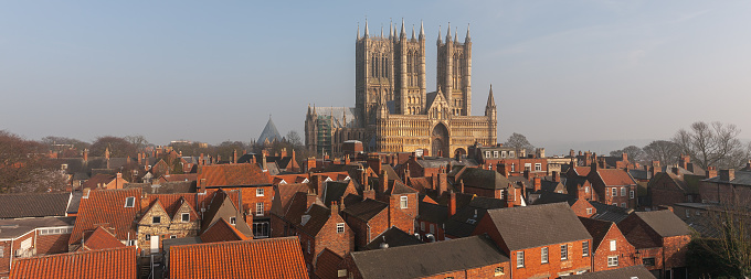 Red brick and red tiled roofs of the old town stand in front of pale yellow stone of Lincoln Cathedral  which rises up behind against a hazy blue sky