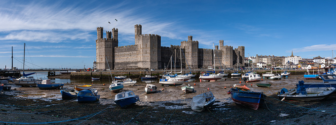 Panorama of Caernarfon Castle in Wales with many boats resting on the muddy bed of the estuary and a bright blue sky with wispy white clouds
