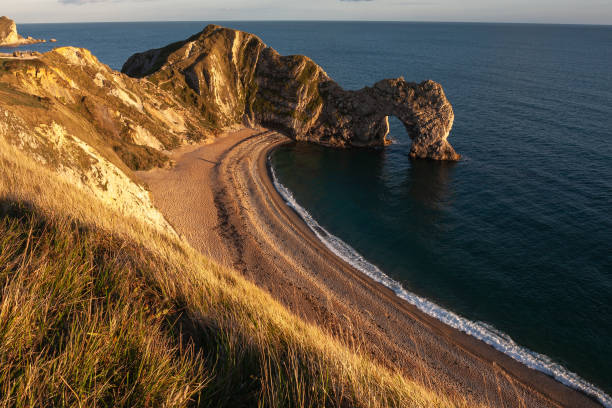 The natural arch of Durdle Door  near Lulworth Cove, seen from the cliffs above with a curve of sand forming the shoreline, all lit by the evening sun