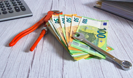 A stack of banknotes with a wrench and a pipe wrench on a wooden background, horizontal