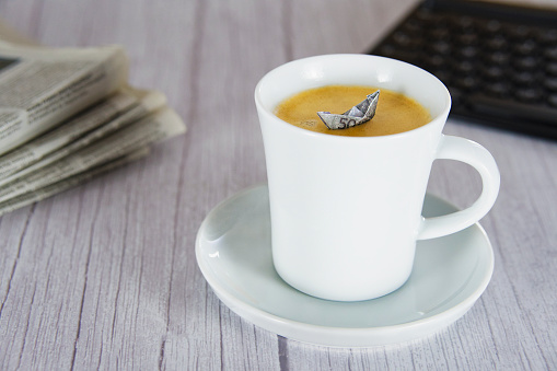 Coffee mug in an oblique view with a paper boat in it, on a wooden background, keyboard and newspaper in the background, horizontal
