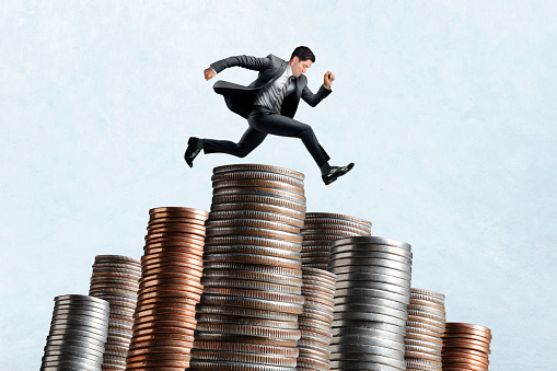 A businessman leaps over a stacks of coins isolated on a light blue background