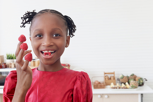 African girl in pink dress with curly hair braid African hairstyle eating ripe fresh raspberries from fingers. Happy kid eating fruit and spending time in kitchen. Cute child in lovely family.