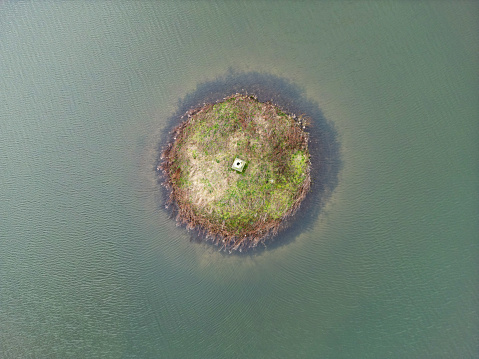 Drone top down view of a small, round aquatic bird sanctuary seen on a fresh water lake in the UK. A central large ceramic ornament can be seen in the middle of the island.