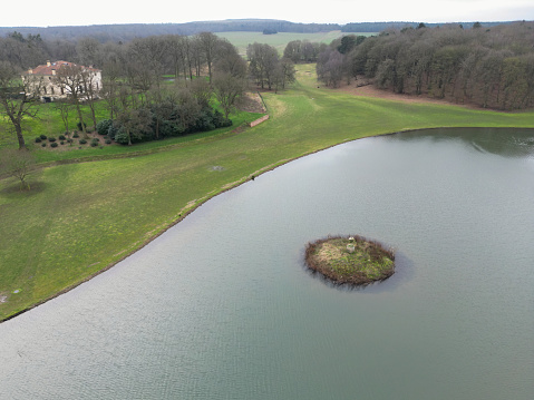 Drone view of a very small, concentric aquatic bird island seen in an English, freshwater lake. A large house can be seen nestled in lake winter woodlands.