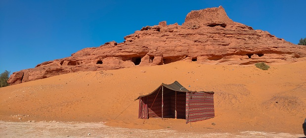 panoramic view of a tent pitched on the sand, and in the background the ruins of Ksar d'ighzer, an ancient stone and red clay castle in the middle of the desert in the town of Timimoun, Algeria