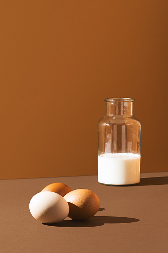Three eggs with milk in glass bottle on brown background, studio shot with sharp shadows and high contrast