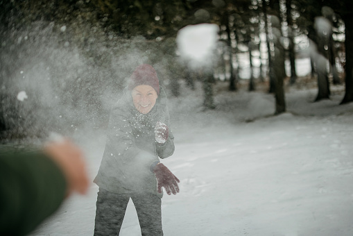 Happy woman having fun while throwing snow towards the camera during winter day in nature.