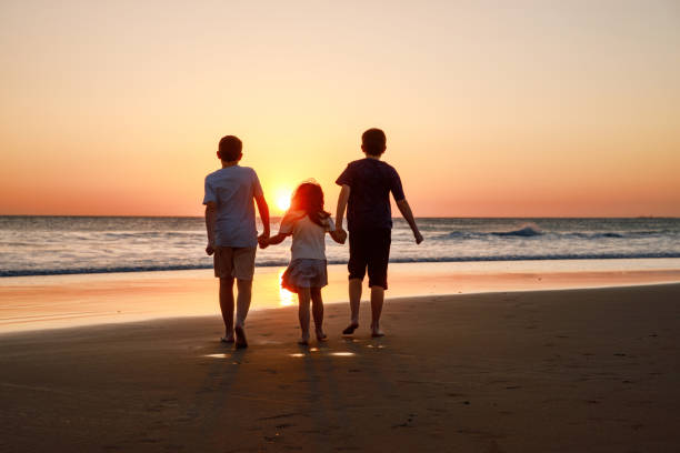 three kids silhouettes running and jumping on beach at sunset. happy family, two school boys and one little preschool girl. siblings having fun together. bonding and family vacation. - silhouette three people beach horizon - fotografias e filmes do acervo