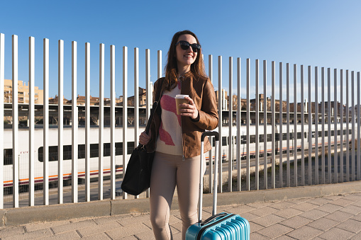 Businesswoman waiting outside the station for the arrival of the train to board. She is carrying a blue suitcase and drinking coffee.