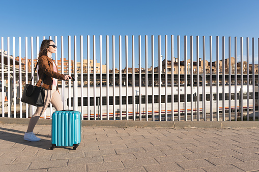 General shot of young woman with blue suitcase walking in the street around the train station. Travel concept.
