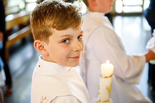 Little kid boy receiving his first holy communion. Happy child holding Christening candle. Tradition in catholic curch. Kid in a white traditional gown in a church near altar