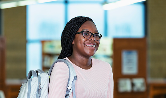 Headshot of a teenage girl with eyeglasses and braces, walking in a library, looking toward the camera with a sideways glance. She is 16 years old, multiracial, black and Hispanic.