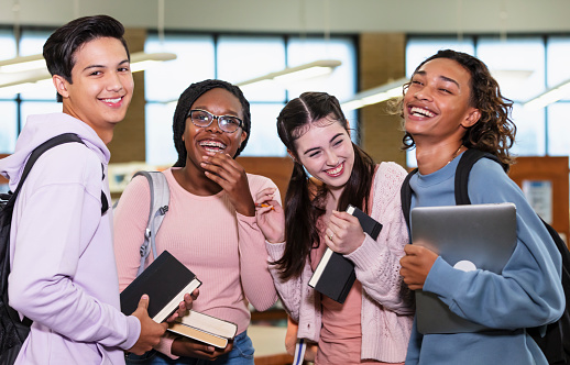A multiracial group of four high school students in a school library carrying backpacks, books and a laptop computer. They are standing together, smiling and laughing at the camera.