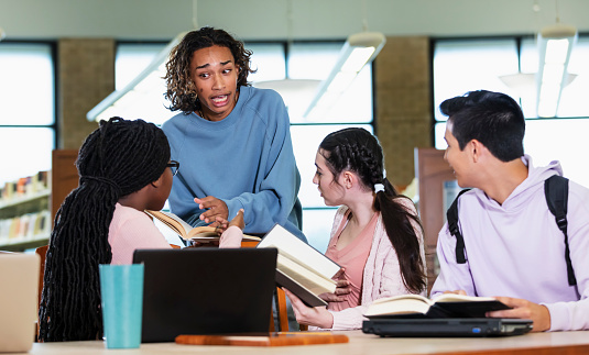 A multiracial group of four high school students studying in a library, working on a school project together. One of the boys, mixed race Black and Hispanic, is standing behind the girls who are sitting at a table. He is talking with a serious expression, having a debate with one of the girls.