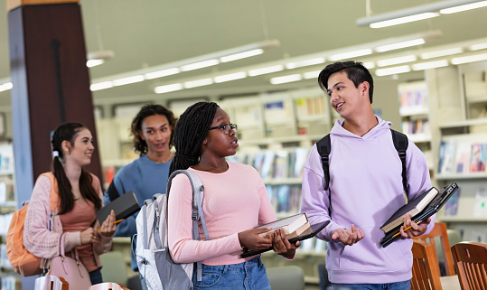 A multiracial group of four high school students carrying backpacks, laptops and textbooks, walking in a library, getting ready to study. They are teenagers, 15 and 16 years old. The focus is on the two friends conversing in the foreground. The girl is black and Hispanic, and the boy is Asian and white.
