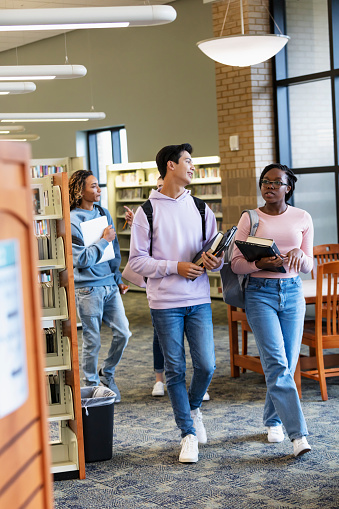 A multiracial group of high school students carrying backpacks, laptops and textbooks, conversing as they walk through a library, getting ready to study.