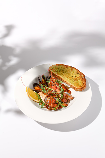 SautÃ©ed shellfish medley with ciabatta toast, side view on white plate with leaf shadow art.