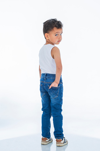 Latin boy with dark skin and curly hair is inside a photography studio where he models a children's clothing line while enjoying the moment and looking at the camera.