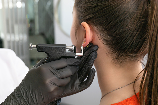 A doctor in black medical gloves pierces the ears of a beautiful young woman in the medical office using a safe black piercing gun and medical earrings. High quality photo