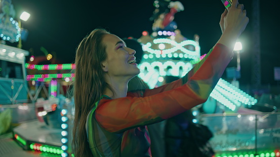 Happy girl recording selfie video at amusement park. Smiling teenager have fun streaming in social media using mobile phone at neon illuminated funfair.