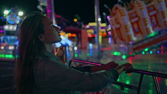 Amazed girl watching funfair carousel closeup. Dreamy young woman resting neon amusement park. Gorgeous model in vibrant festival lights. Relaxed teenager waiting friends enjoying weekend nightlife.