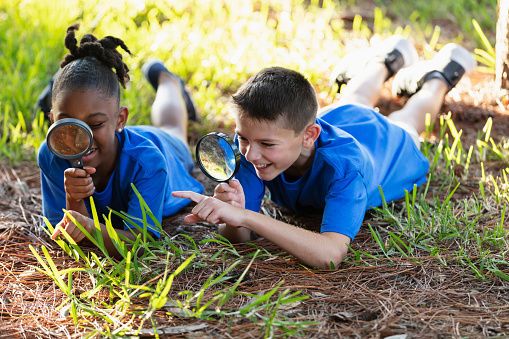 Two multiracial children on a field trip at a park, lying on the grass with magnifying glasses, exploring the environment, examining plant life and insects on the ground.